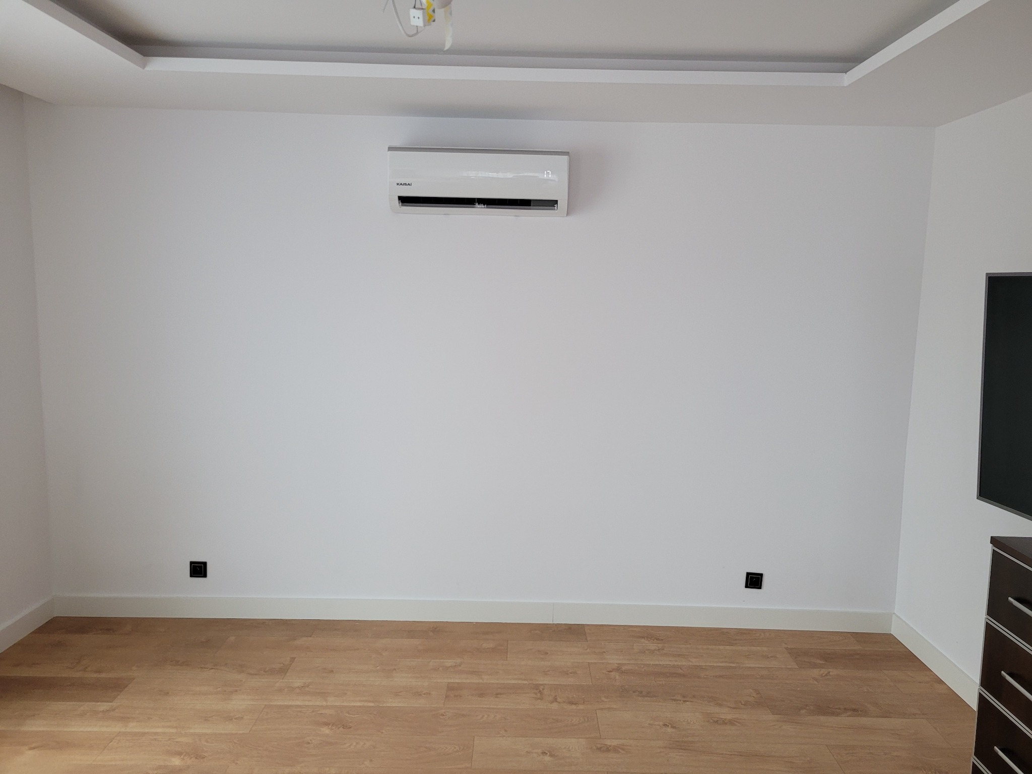 Wnętrze pokoju z zamontowanym klimatyzatorem Kaisai na białej ścianie, widoczne gniazdka elektryczne i drewniana podłoga.