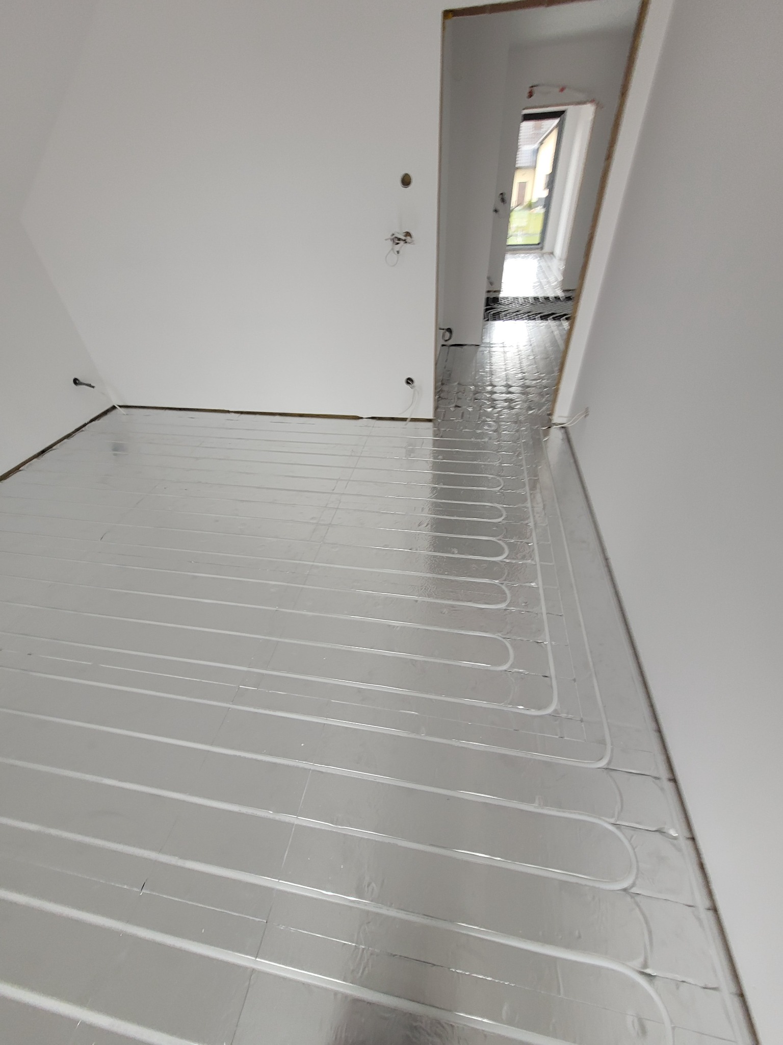 Białe, surowe wnętrze w trakcie instalacji ogrzewania podłogowego; widoczne rury grzewcze ułożone na srebrnej folii izolacyjnej, przygotowane do zalania wylewką.