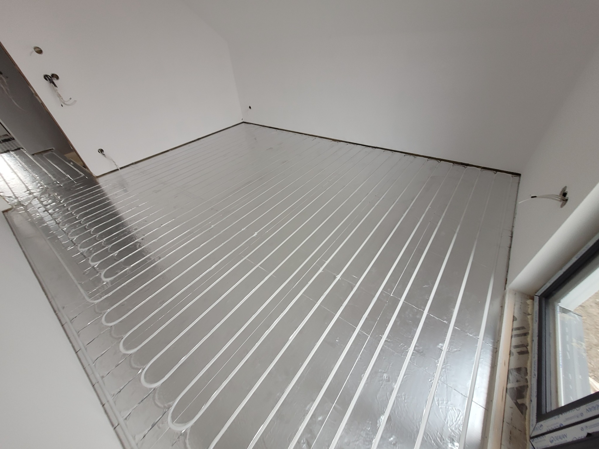 Białe rurki ogrzewania podłogowego ułożone w równych odstępach na srebrnej folii izolacyjnej w nowym budynku, widok z góry na fragment pomieszczenia z oknem.