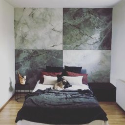 Tinta - Sypialnia z dekoracyjną ścianą imitującą marmur w odcieniach szarości i zieleni, podzieloną na cztery kwadraty, z psem rasy mops leżącym na łóżku przykrytym ciemnym pledem.