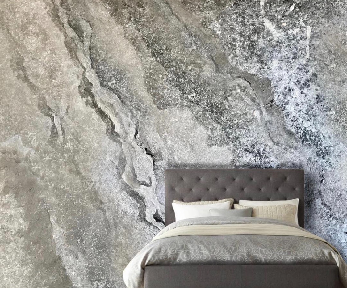 Sypialnia z dekoracyjną ścianą imitującą szary kamień z wyraźnymi żyłami i nieregularną fakturą, zagłówek łóżka w kolorze popielatym.