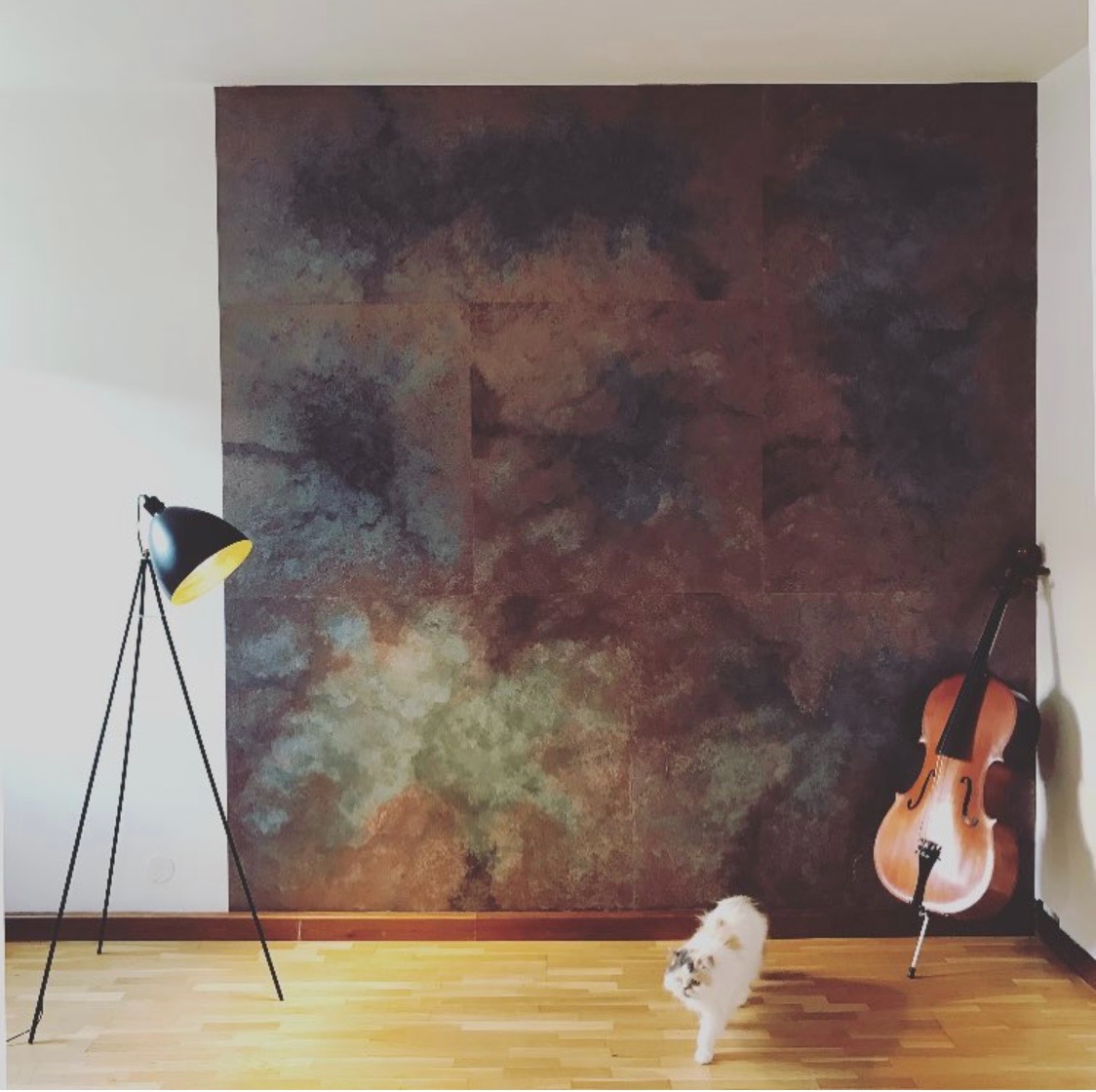 Artystycznie pomalowana ściana w odcieniach brązu i błękitu, przypominająca abstrakcyjny krajobraz, z wiolonczelą opartą o ścianę, czarną lampą na trójnogu i kotem wchodzącym w kadr.