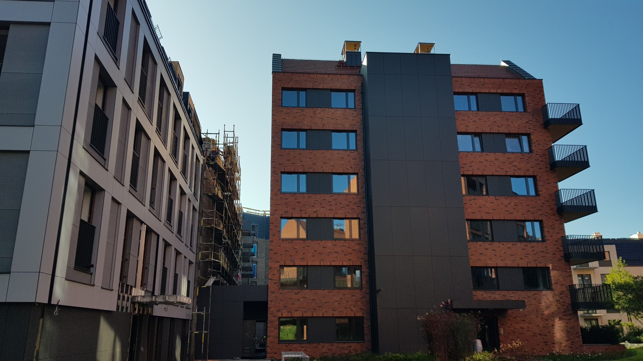 Nowoczesny budynek z elewacją z cegły klinkierowej i szarą okładziną, z balkonami z metalowymi barierkami, obok budynek w trakcie wykańczania elewacji wentylowanej z użyciem rusztowań.