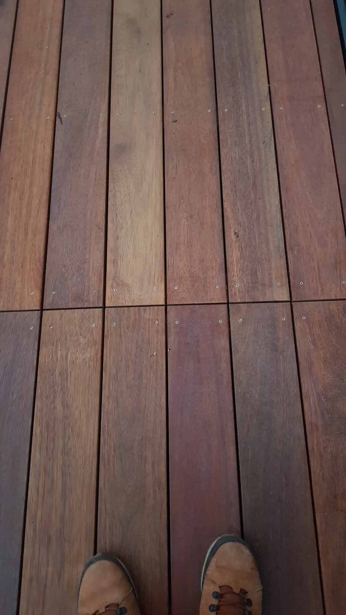 Detale montażu tarasu wentylowanego z drewna egzotycznego: widoczne deski tekowe mocowane na wkręty, z fragmentem buta roboczego w dolnej części kadru.