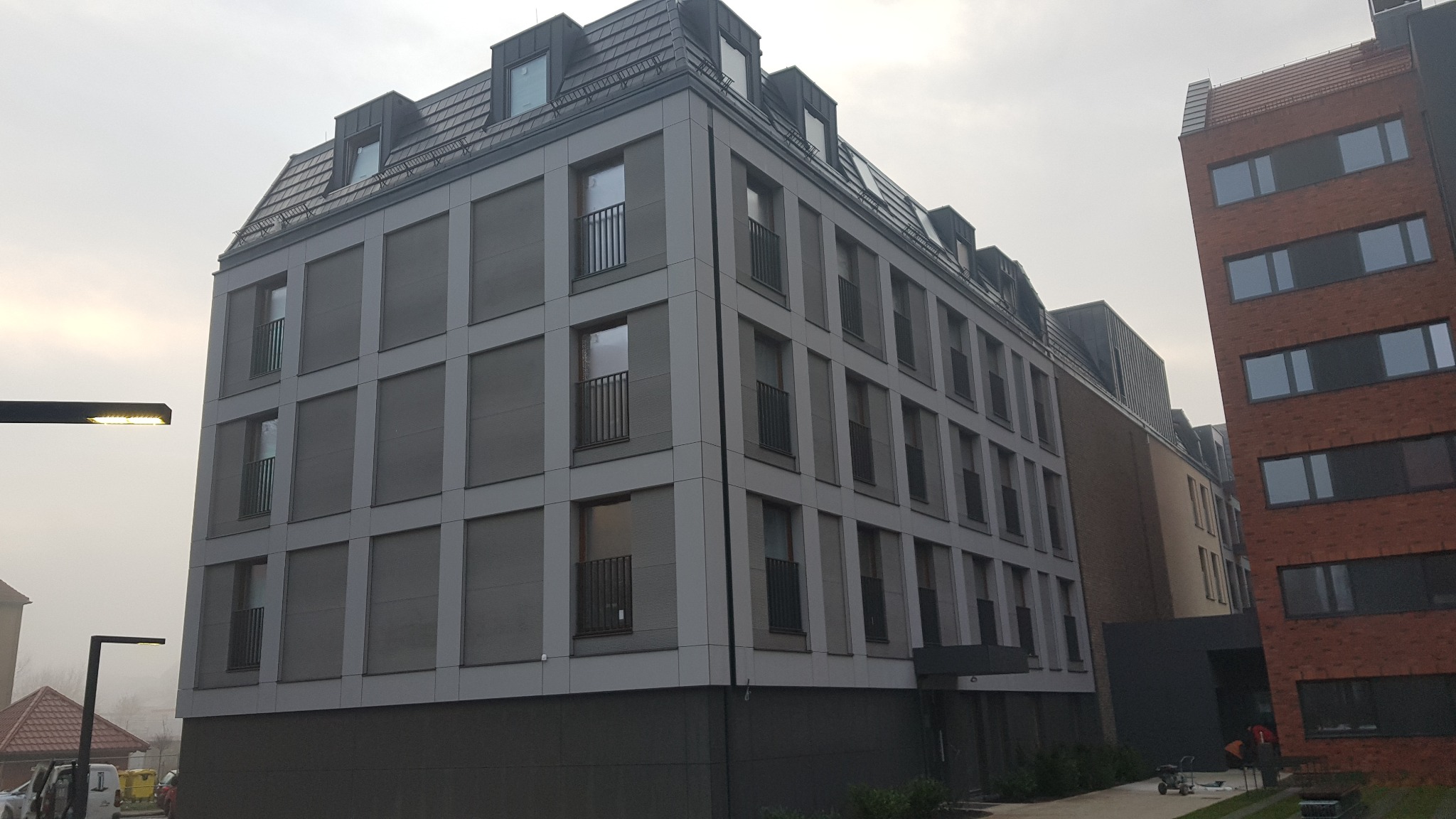 Nowoczesny budynek wielorodzinny z szarej elewacji z elementami pionowych podziałów, małymi balkonami i mansardowym dachem w pochmurny dzień.