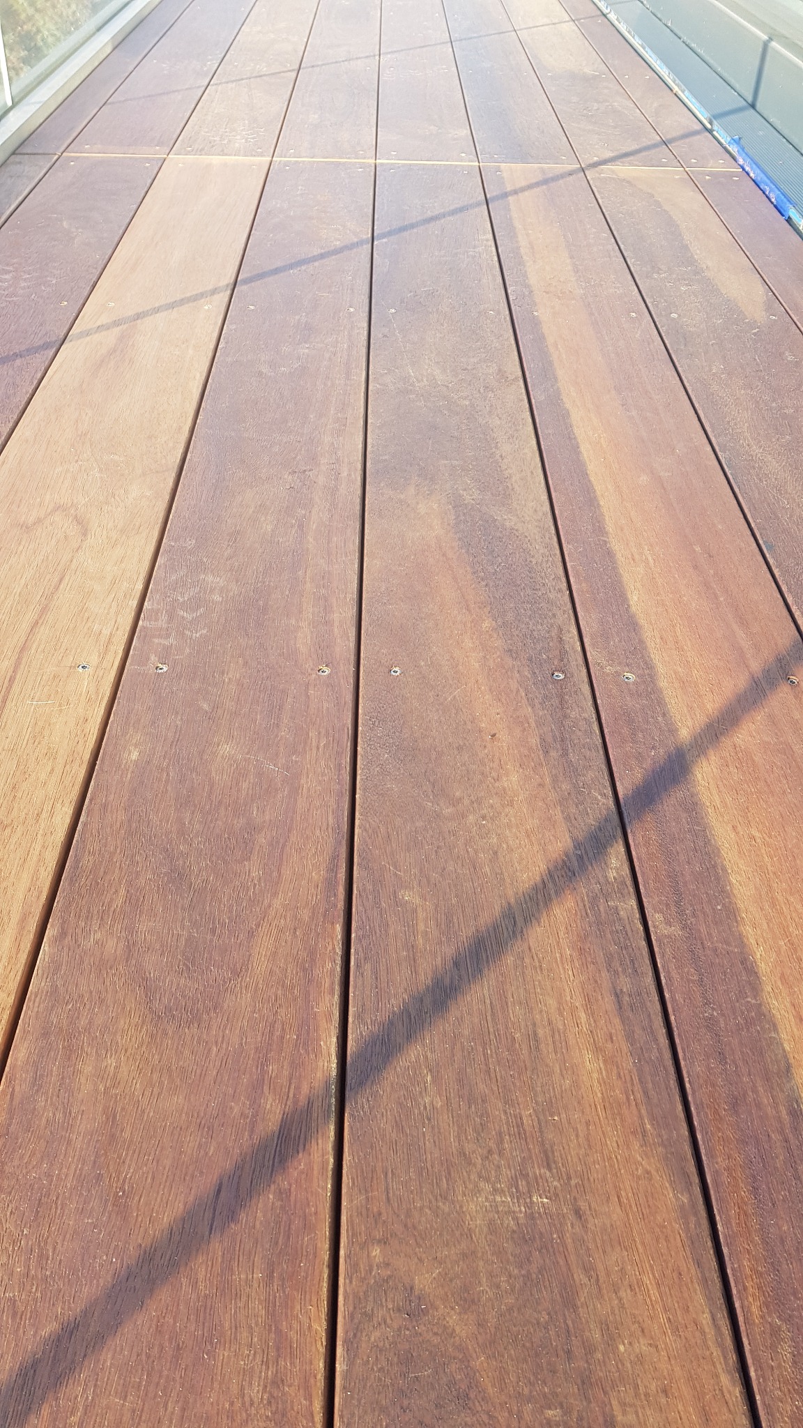 Szczegółowe ujęcie desek tarasowych z egzotycznego drewna tekowego, widoczne śruby mocujące i cień padający na powierzchnię.