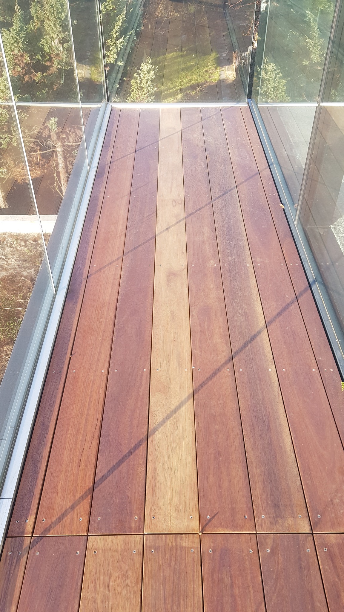 Nowoczesny taras wentylowany z drewna egzotycznego, z widocznymi śrubami mocującymi deski i szklaną balustradą, odbijającą zieleń otoczenia.