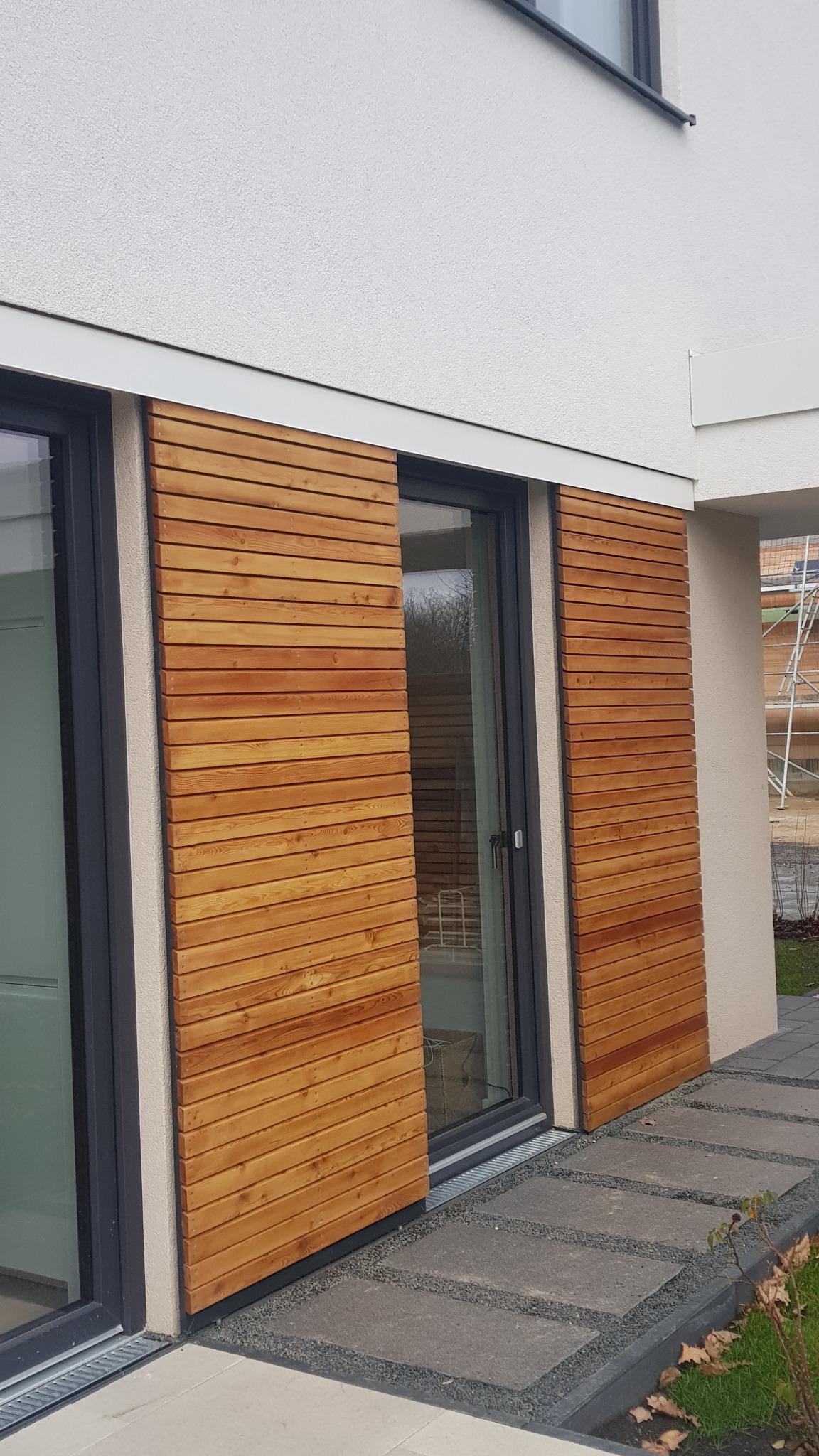 Zewnętrzne rolety przesuwne z drewna modrzewiowego, olejowane, zamontowane na aluminiowej konstrukcji, widoczne na tle białej ściany domu i szarego chodnika.