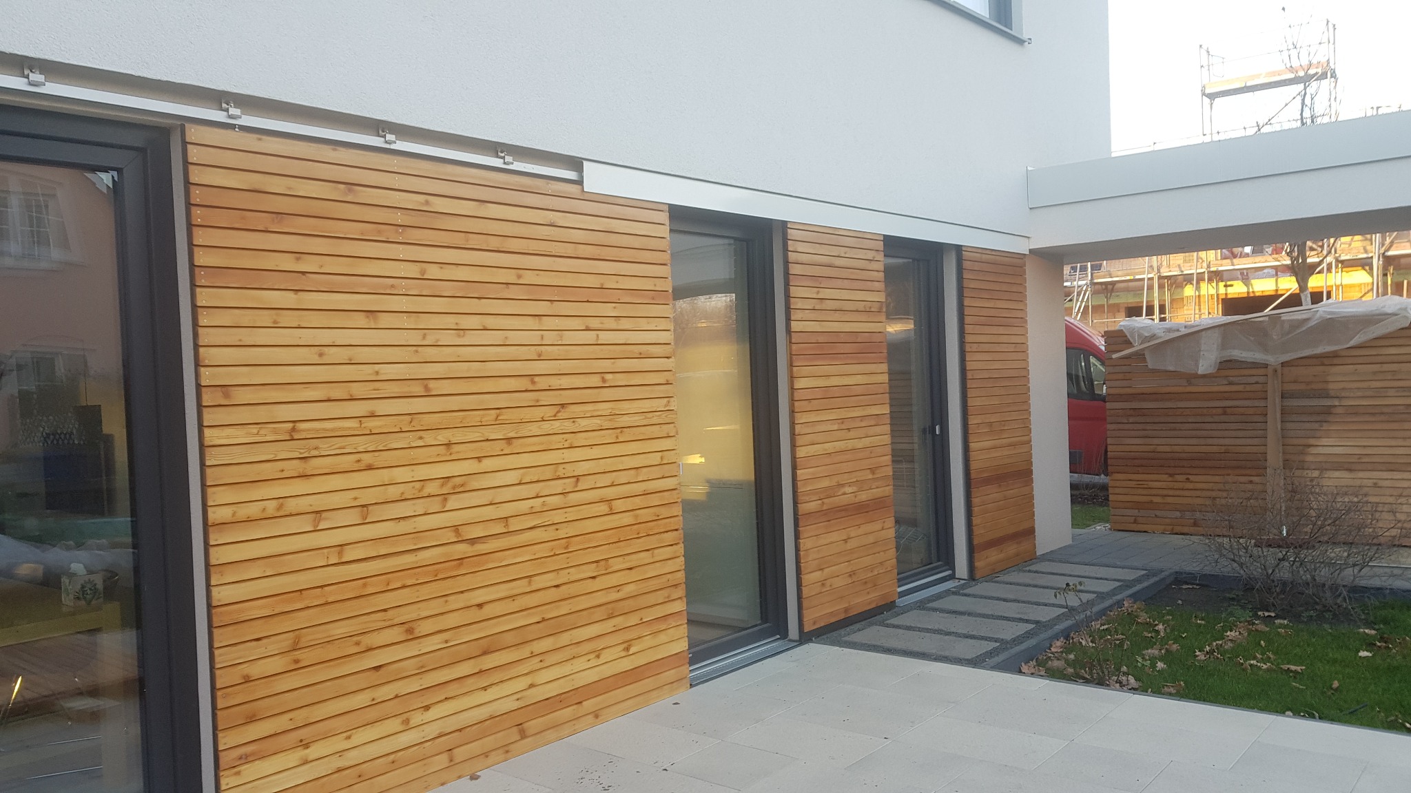 Elewacja budynku z przesuwnymi panelami z modrzewiowego drewna, zamontowanymi na aluminiowej konstrukcji, widoczna szyna prowadząca i przesuwne drzwi.