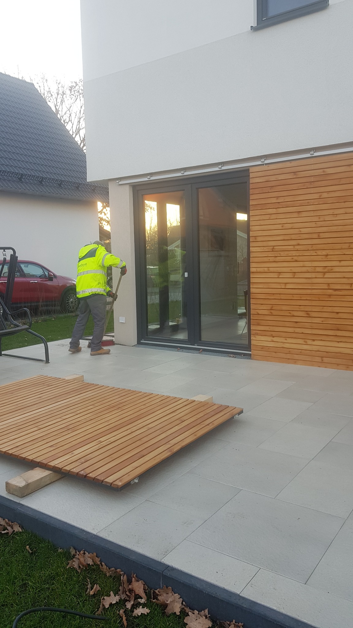 Pracownik w odblaskowej kamizelce zamiata nowo położony taras z szarych płyt betonowych, z częściowo ułożonym drewnianym panelem na podkładkach, przesuwne drzwi tarasowe i drewniana elewacja w tle.