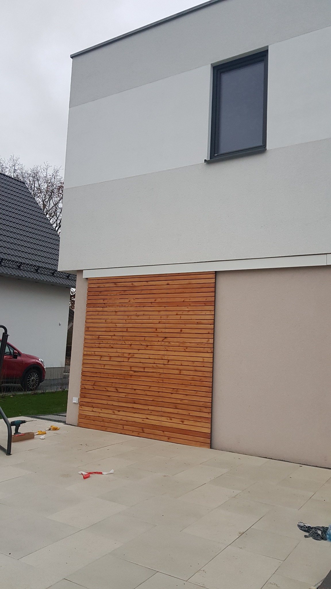 Zewnętrzna ściana budynku z zamontowaną drewnianą, przesuwną osłoną z poziomych desek z jasnego drewna, widoczny fragment tarasu z jasnych płyt i fragment ściany budynku pomalowanej na szaro.