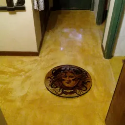 Żółta posadzka żywiczna z wkomponowanym okrągłym elementem dekoracyjnym przedstawiającym głowę Meduzy, widoczna w korytarzu z ciemnymi drzwiami i fragmentami ścian.