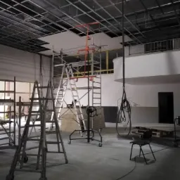 Wnętrze w trakcie prac wykończeniowych, widoczne rusztowania, drabiny, częściowo zamontowany sufit podwieszany i przewody zwisające z góry.