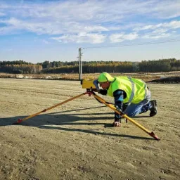 Geodeta w żółtej kamizelce, klęczący przy niwelatorze na terenie budowy, w tle las i słupy wysokiego napięcia.