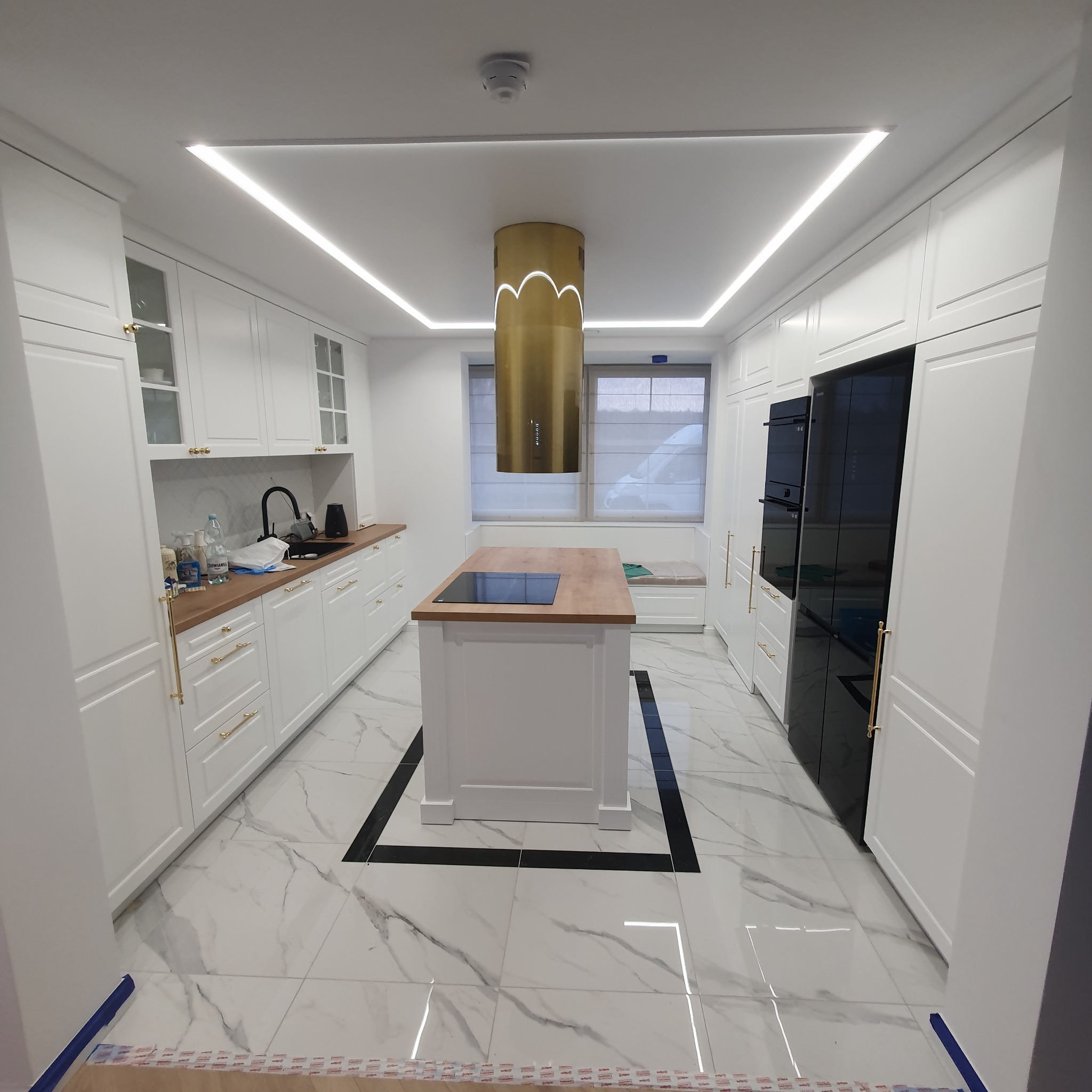 Nowoczesna kuchnia z białymi szafkami, drewnianym blatem, złotym okapem i wyspą na tle marmurowej podłogi z czarnymi akcentami.