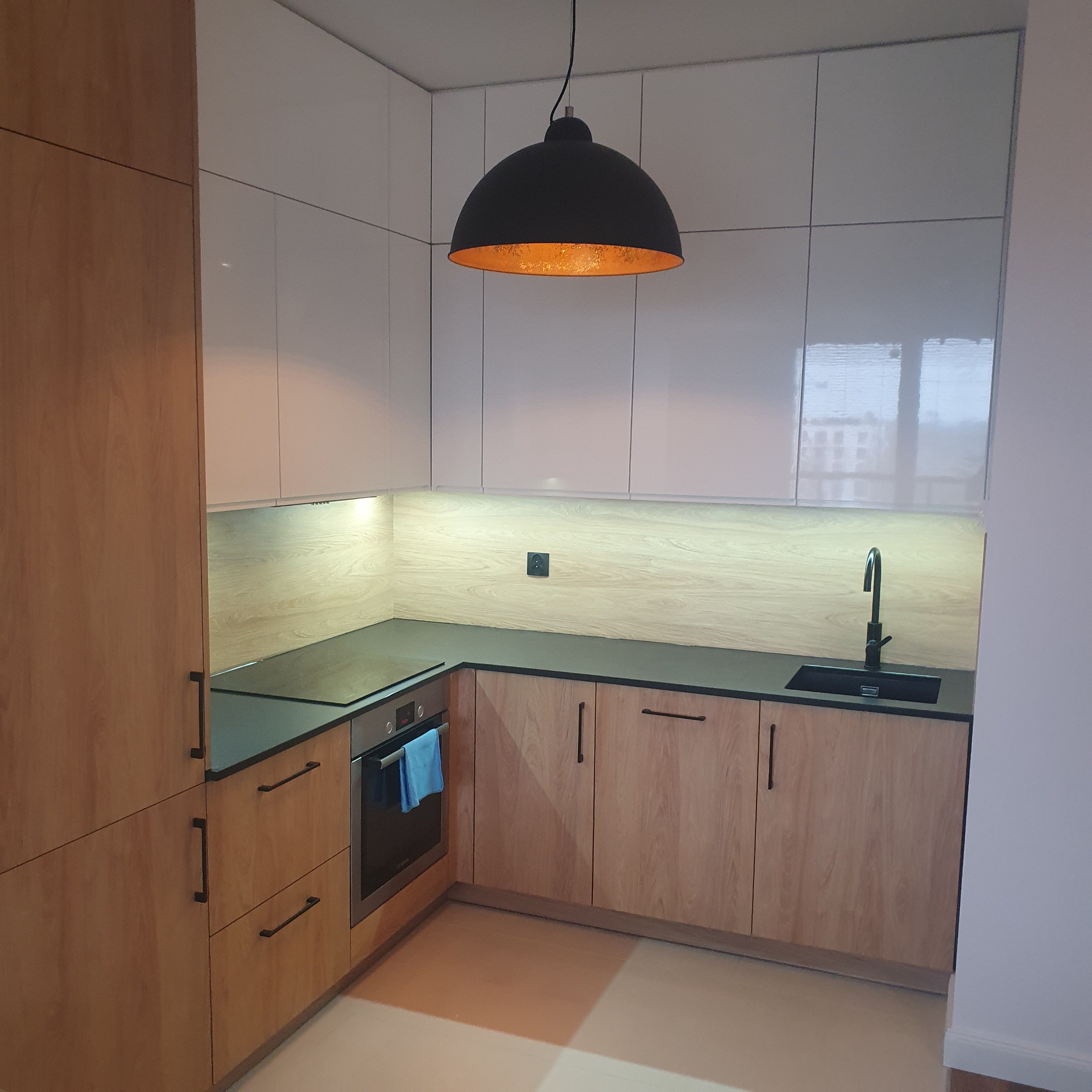 Nowoczesna kuchnia z frontami w kolorze jasnego drewna i białymi szafkami wiszącymi, czarnym blatem i lampą wiszącą w kształcie kopuły z miedzianym wnętrzem. W tle widoczne okno.