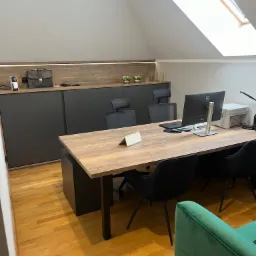 Biuro na poddaszu z drewnianym biurkiem, czarnymi krzesłami i szafką z przesuwnymi drzwiami w kolorze grafitowym, oświetlenie LED pod szafką, okno dachowe.