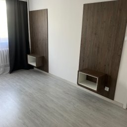 4Mat Meble na wymiar - Dwie pionowe, drewniane panele ścienne z wbudowanymi, kwadratowymi półkami nocnymi z białym wnętrzem, zamontowane na białej ścianie w sypialni z szarymi panelami podłogowymi i czarnymi zasłonami.