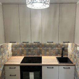 4Mat Meble na wymiar - Nowoczesna kuchnia z kremowymi szafkami, drewnianym blatem, wzorzystymi płytkami ściennymi i czarną armaturą, oświetlona dekoracyjną lampą sufitową.