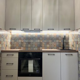 4Mat Meble na wymiar - Jasnoszara zabudowa kuchenna z czarnymi uchwytami, wzorzyste płytki nad blatem i czarna bateria.