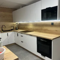 Nowoczesna kuchnia z białymi frontami i drewnianym blatem, czarna armatura i sprzęt AGD marki Bosch, oświetlenie LED pod szafkami wiszącymi.