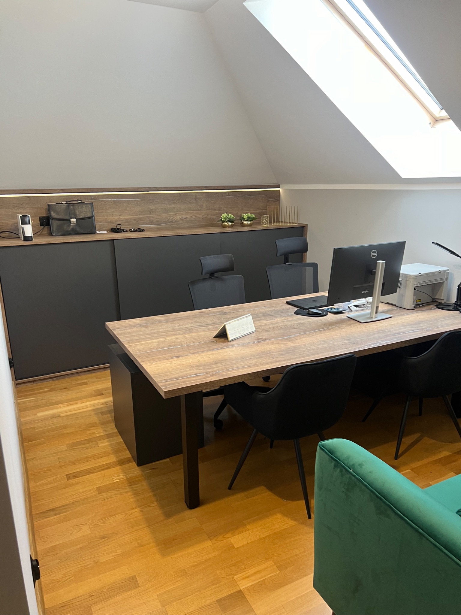 Biuro na poddaszu z drewnianym biurkiem, czarnymi krzesłami i szafką z przesuwnymi drzwiami w kolorze grafitowym, oświetlenie LED pod szafką, okno dachowe.