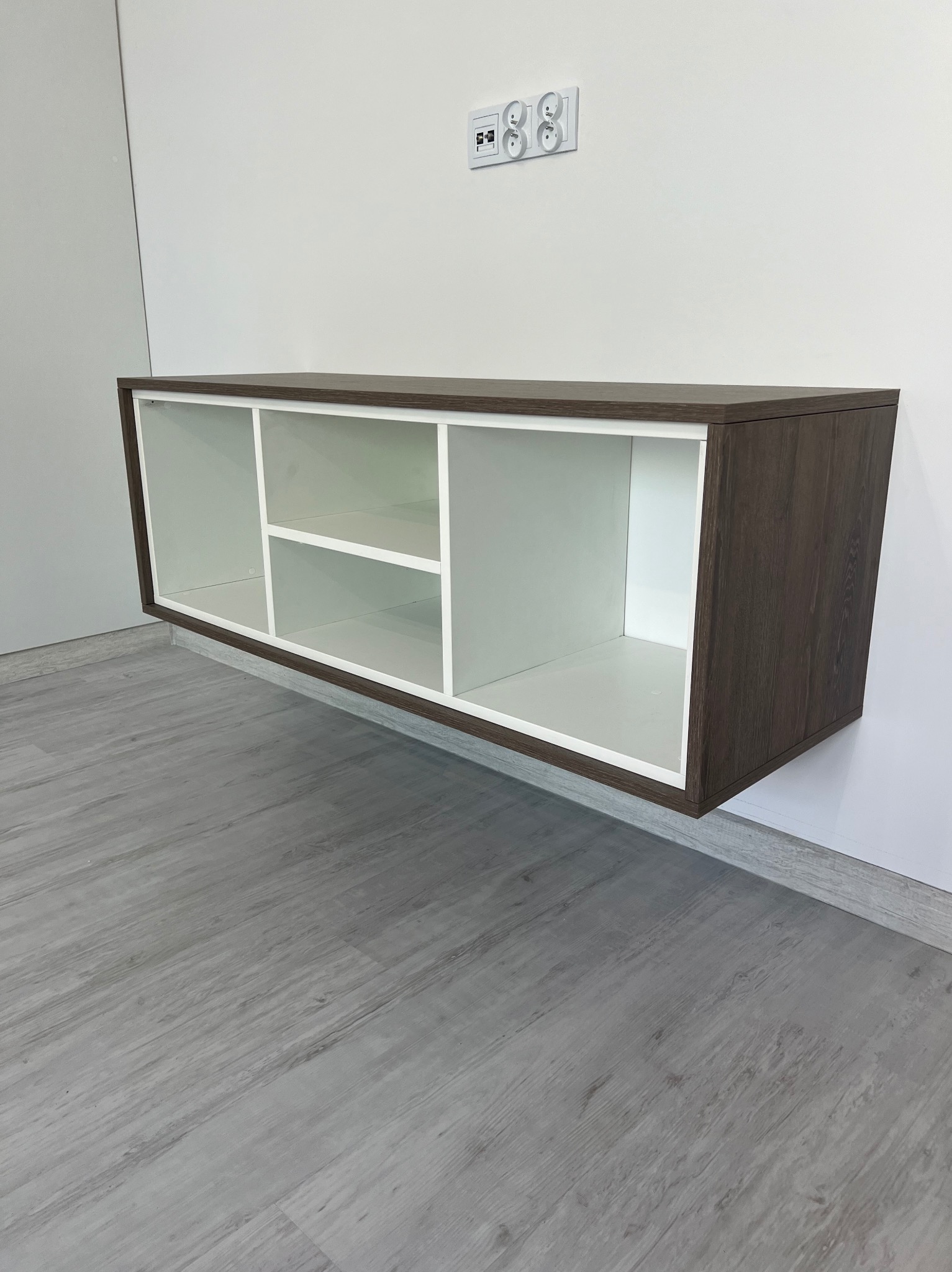 Półka wisząca w kolorze ciemnego drewna z białymi wnętrzami, zamontowana na białej ścianie nad szarą podłogą imitującą drewno, widoczne gniazdka elektryczne nad meblem.