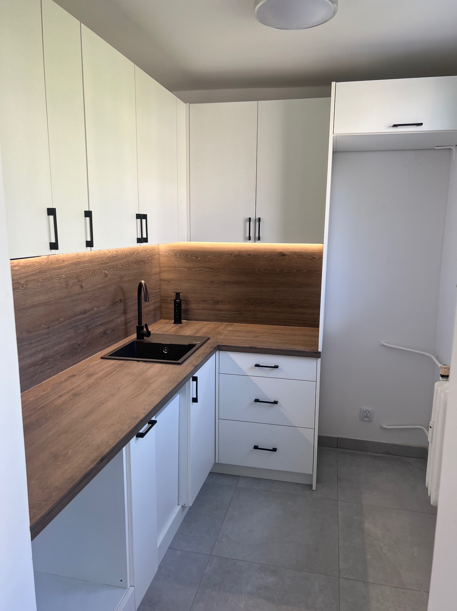 Nowoczesna kuchnia na wymiar w Dębnie z białymi szafkami i drewnopodobnym blatem, czarny zlew i bateria, oświetlenie LED pod szafkami wiszącymi.