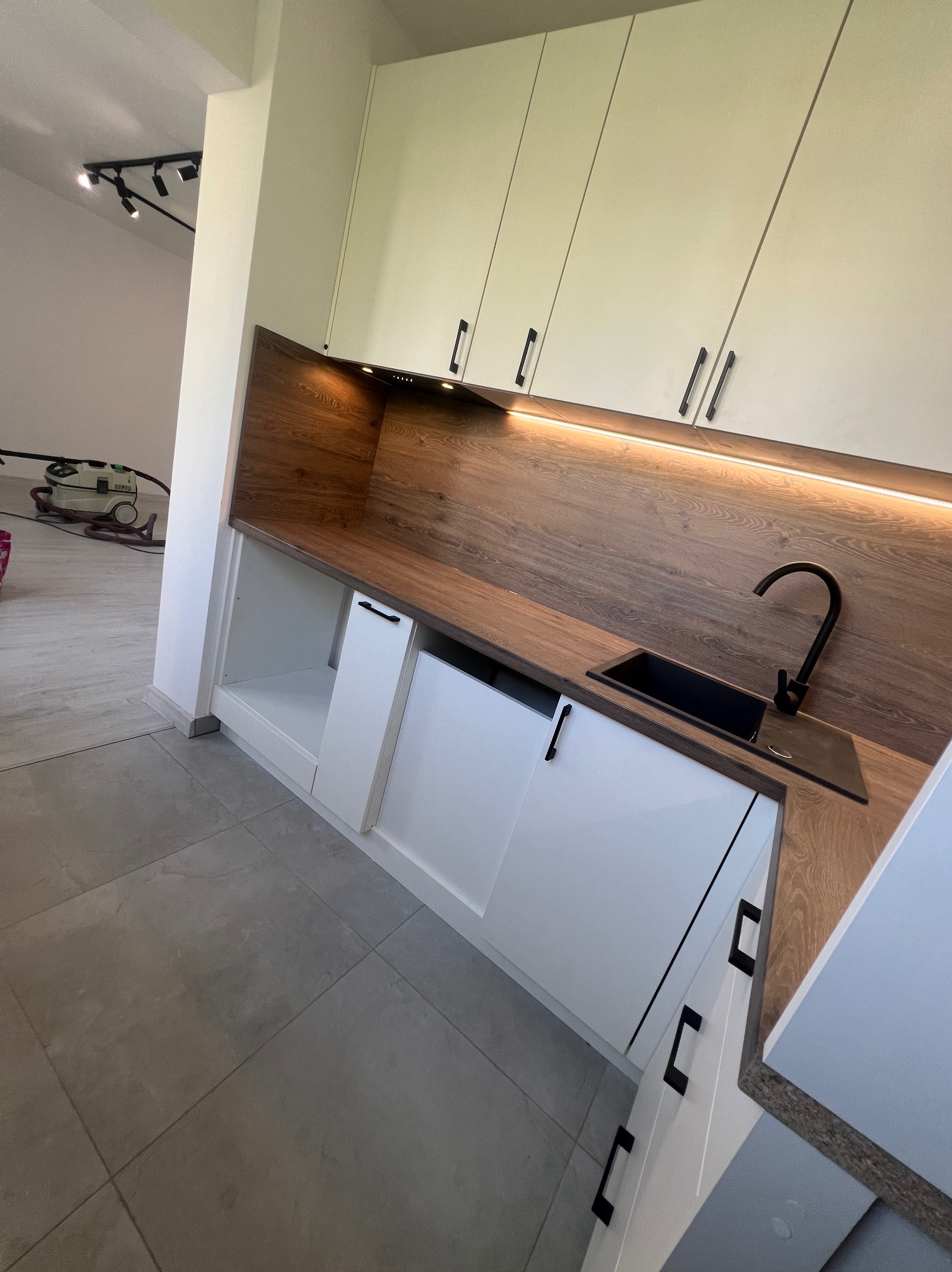 Nowoczesna kuchnia z białymi szafkami, drewnianym blatem i podświetleniem LED, czarna armatura i zlewozmywak, na podłodze szare płytki, w tle widoczny odkurzacz przemysłowy.