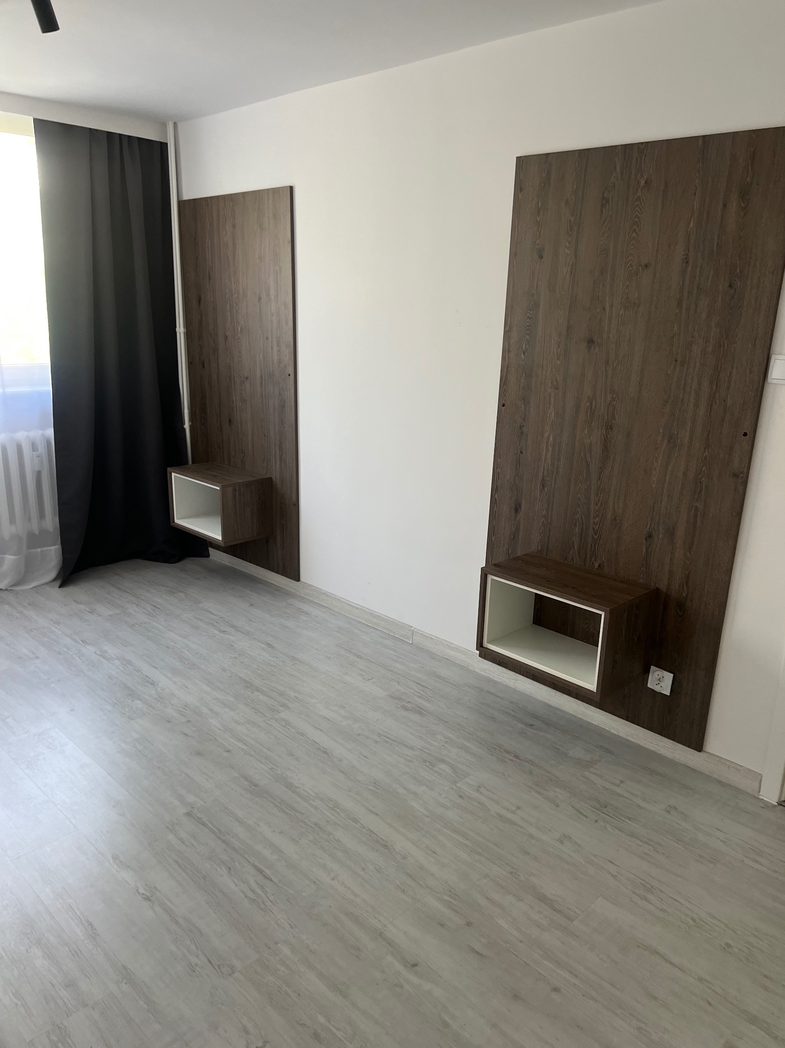 Dwie pionowe, drewniane panele ścienne z wbudowanymi, kwadratowymi półkami nocnymi z białym wnętrzem, zamontowane na białej ścianie w sypialni z szarymi panelami podłogowymi i czarnymi zasłonami.