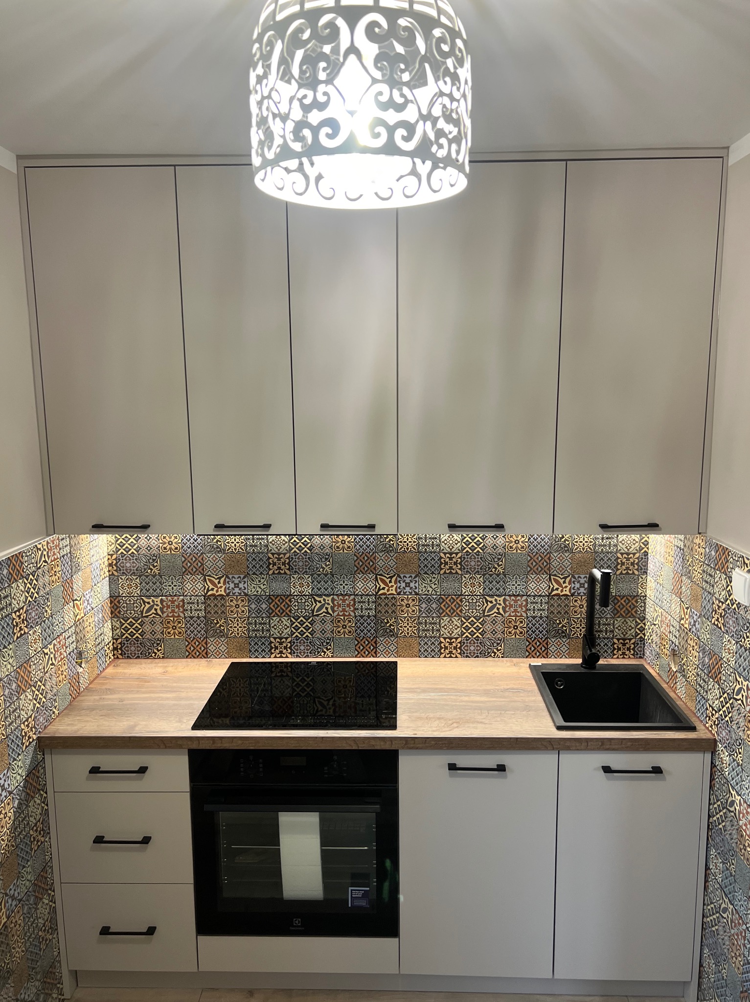 Nowoczesna kuchnia z kremowymi szafkami, drewnianym blatem, wzorzystymi płytkami ściennymi i czarną armaturą, oświetlona dekoracyjną lampą sufitową.