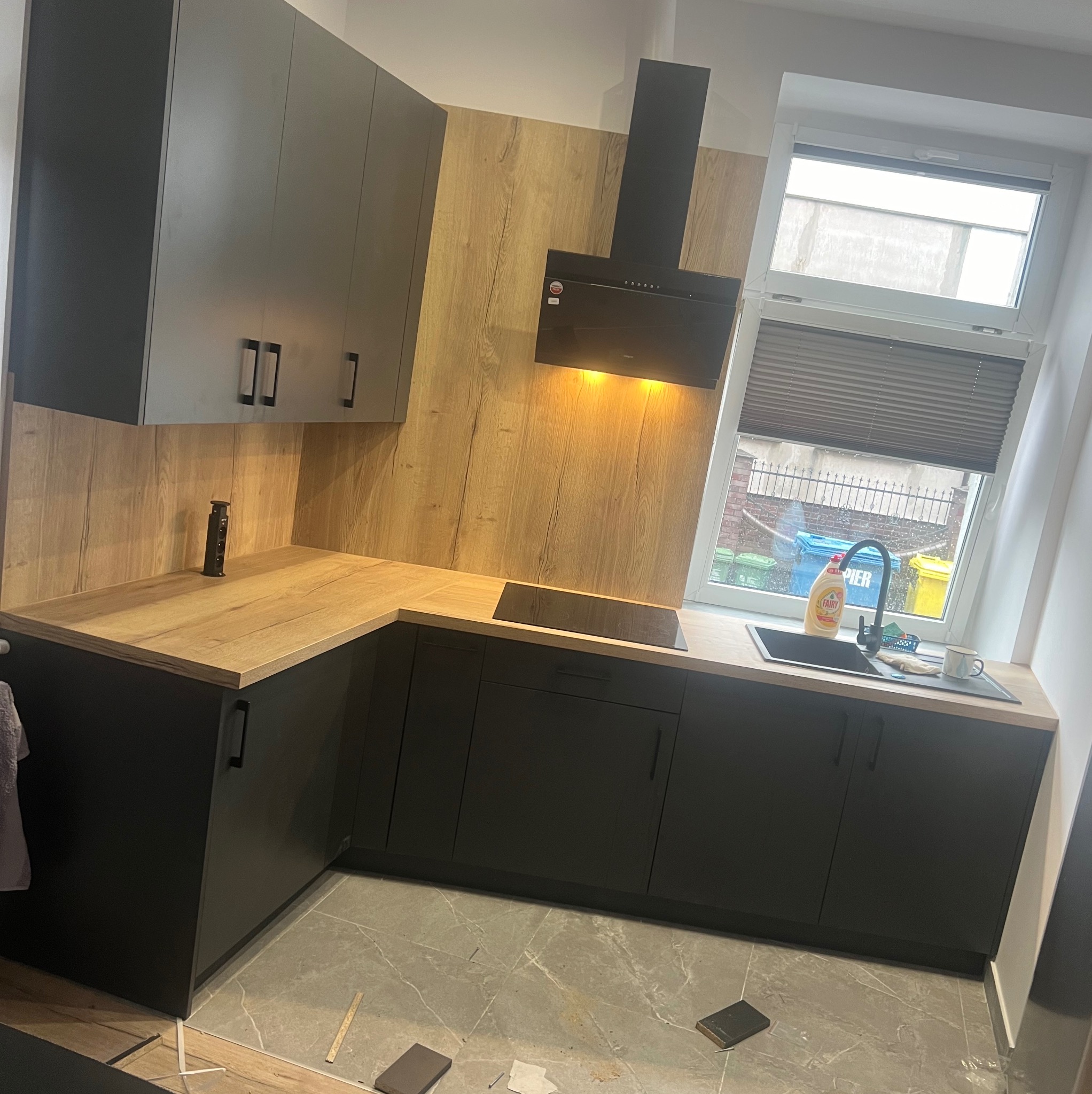 Nowoczesna kuchnia z szarymi szafkami, drewnianym blatem i okładziną ścienną, czarnym okapem i płytą indukcyjną, widok na podwórko z koszami na śmieci przez okno.