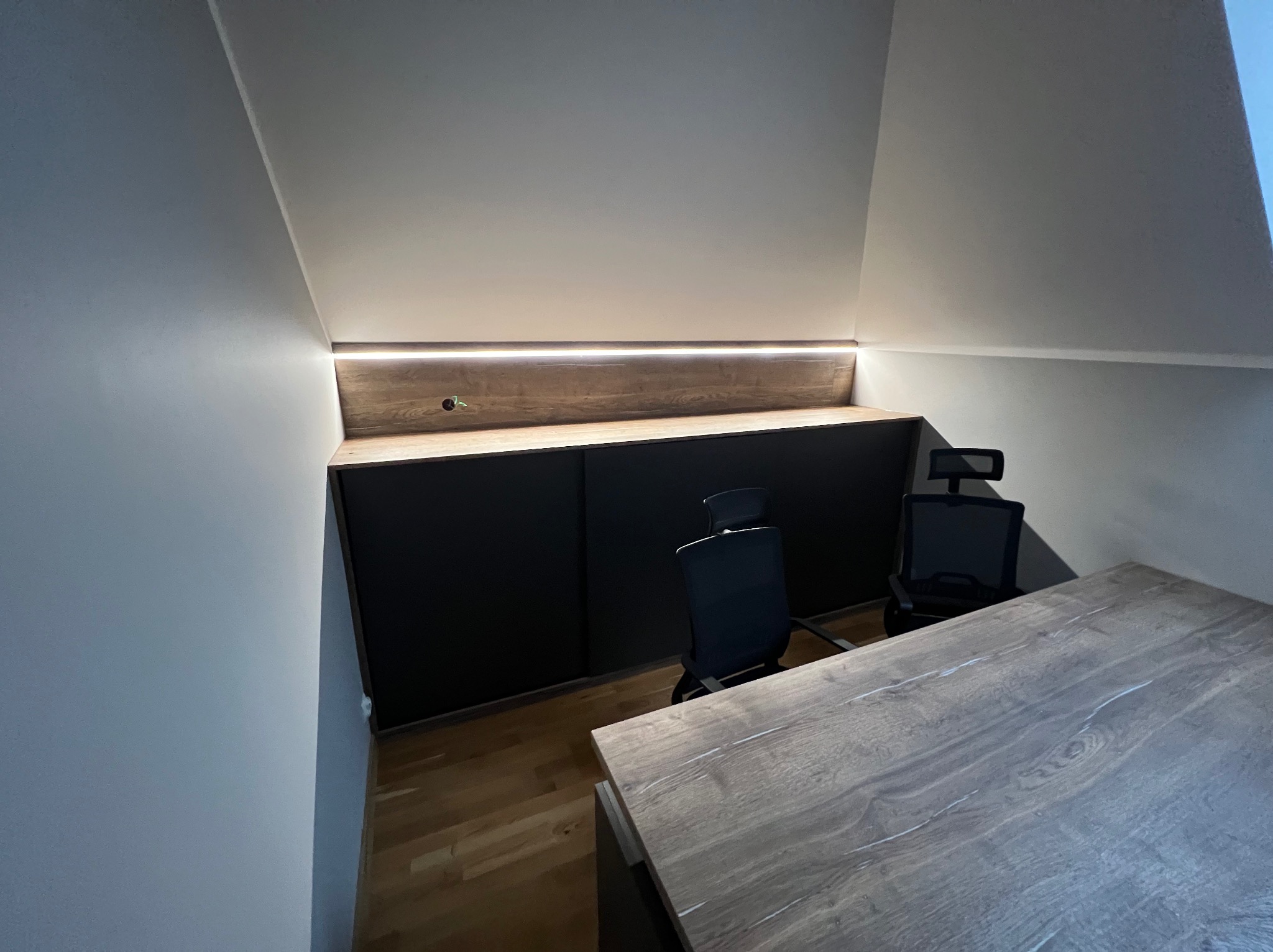 Szafa wnękowa z czarnymi frontami i drewnianym blatem, podświetlona listwą LED, wkomponowana w skos dachu, z biurkiem i krzesłami biurowymi.