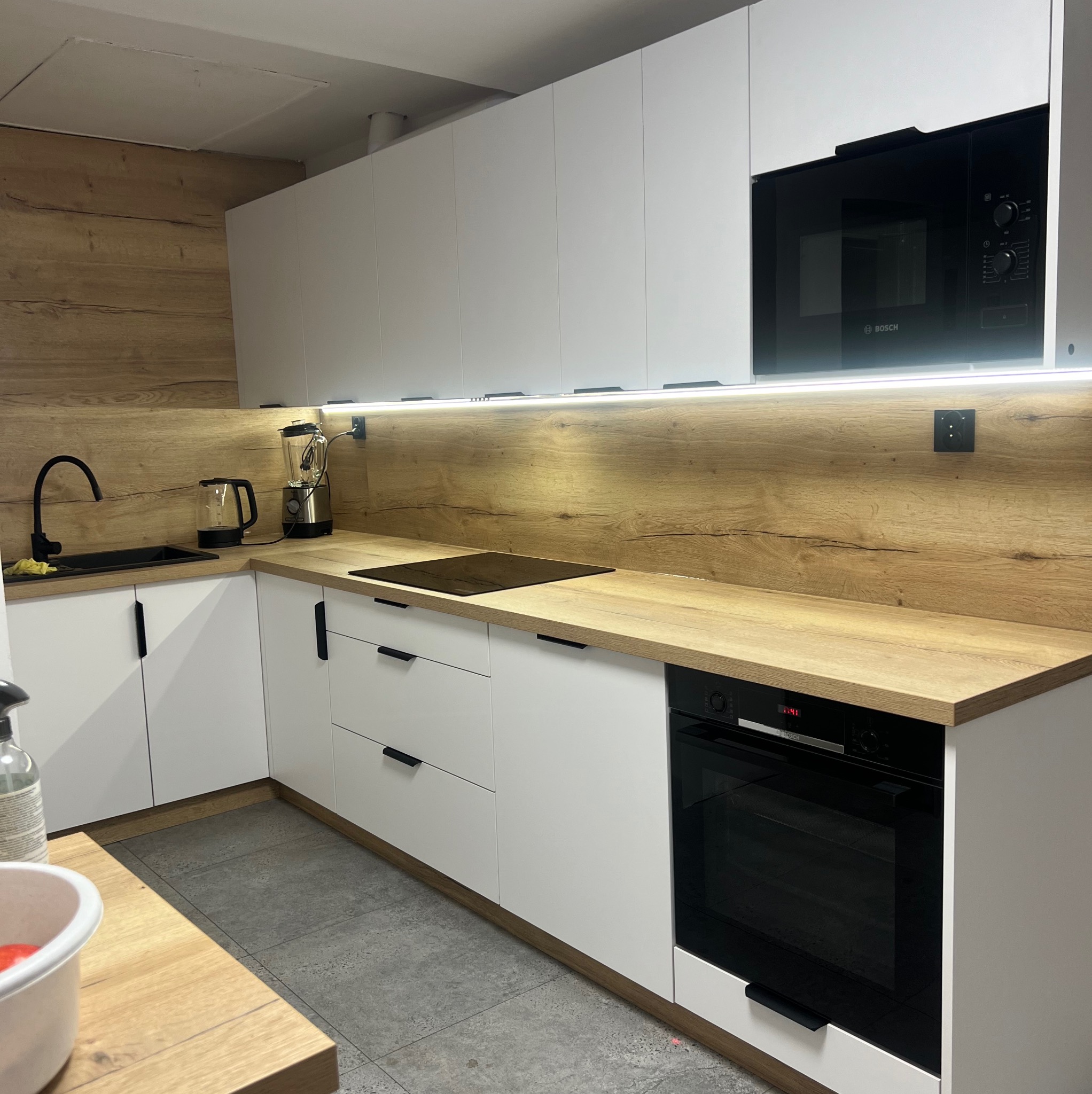 Nowoczesna kuchnia z białymi frontami i drewnianym blatem, czarna armatura i sprzęt AGD marki Bosch, oświetlenie LED pod szafkami wiszącymi.