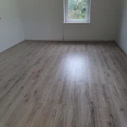 Jasny, dębowy parkiet ułożony w pustym pokoju z białymi ścianami i oknem z widokiem na zieleń.