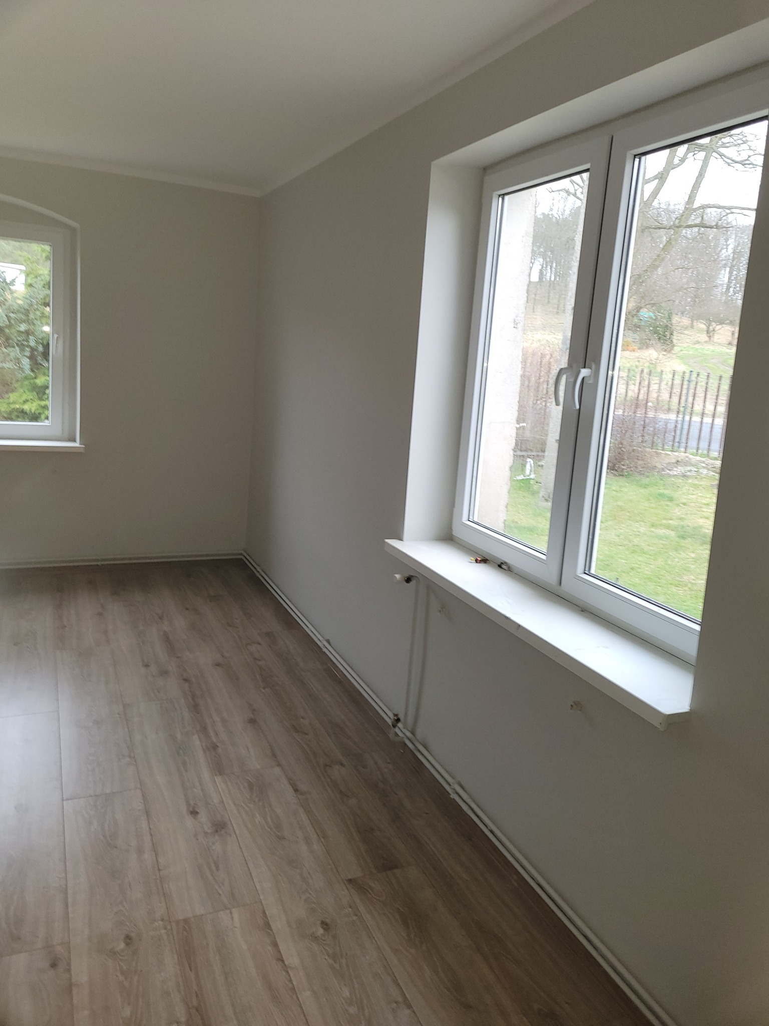 Pusty pokój po remoncie z panelami podłogowymi w odcieniach brązu i beżu, świeżo pomalowanymi ścianami na jasny szaro-beżowy kolor oraz dwoma oknami PCV z białymi parapetami.