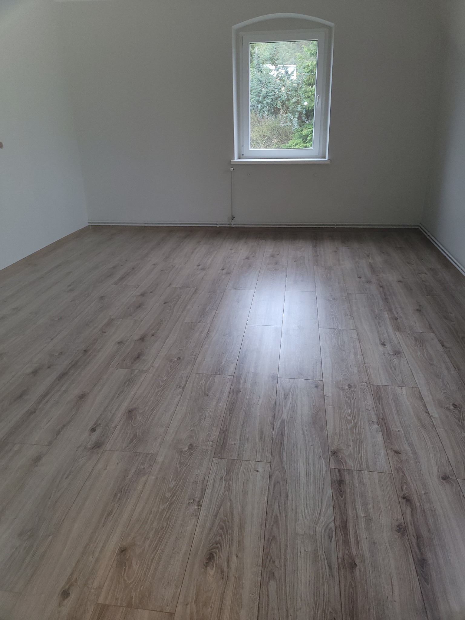 Jasny, dębowy parkiet ułożony w pustym pokoju z białymi ścianami i oknem z widokiem na zieleń.