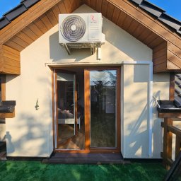 Instalacje Budynkowe Klima4You Mirosław Oleszczuk - Zewnętrzny agregat klimatyzacji zamontowany nad drzwiami tarasowymi domu z drewnianą podbitką dachową i sztuczną trawą na tarasie.