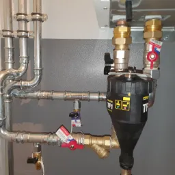 Szczegółowe ujęcie instalacji centralnego ogrzewania z filtrem magnetycznym TF1, zaworami kulowymi z czerwonymi rączkami i rurami ze stali ocynkowanej, zamontowanej na szarej ścianie.