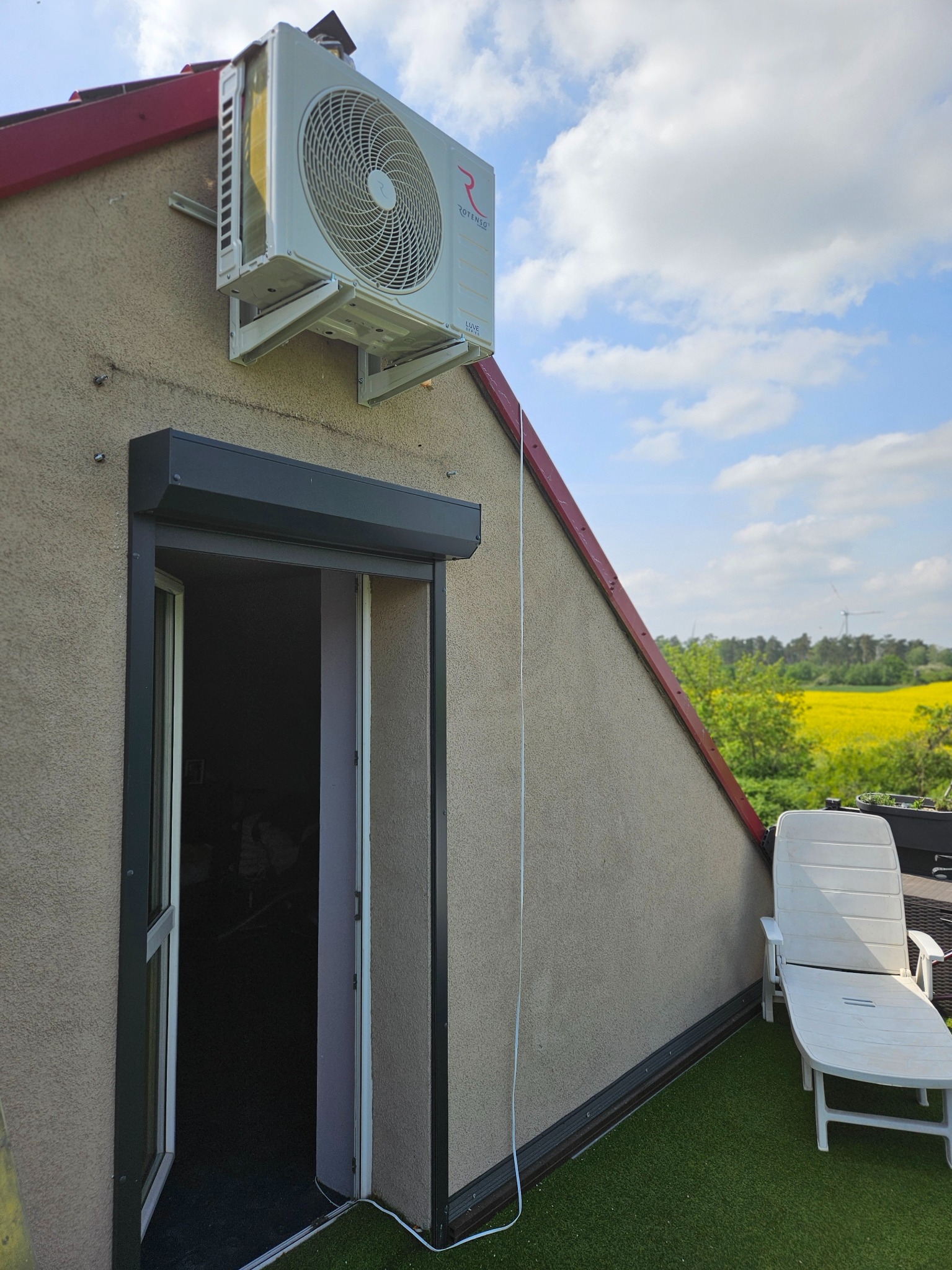 Zewnętrzna jednostka klimatyzacji zamontowana na ścianie szczytowej budynku z oknem roletowym, widok na żółte pole rzepaku w tle.