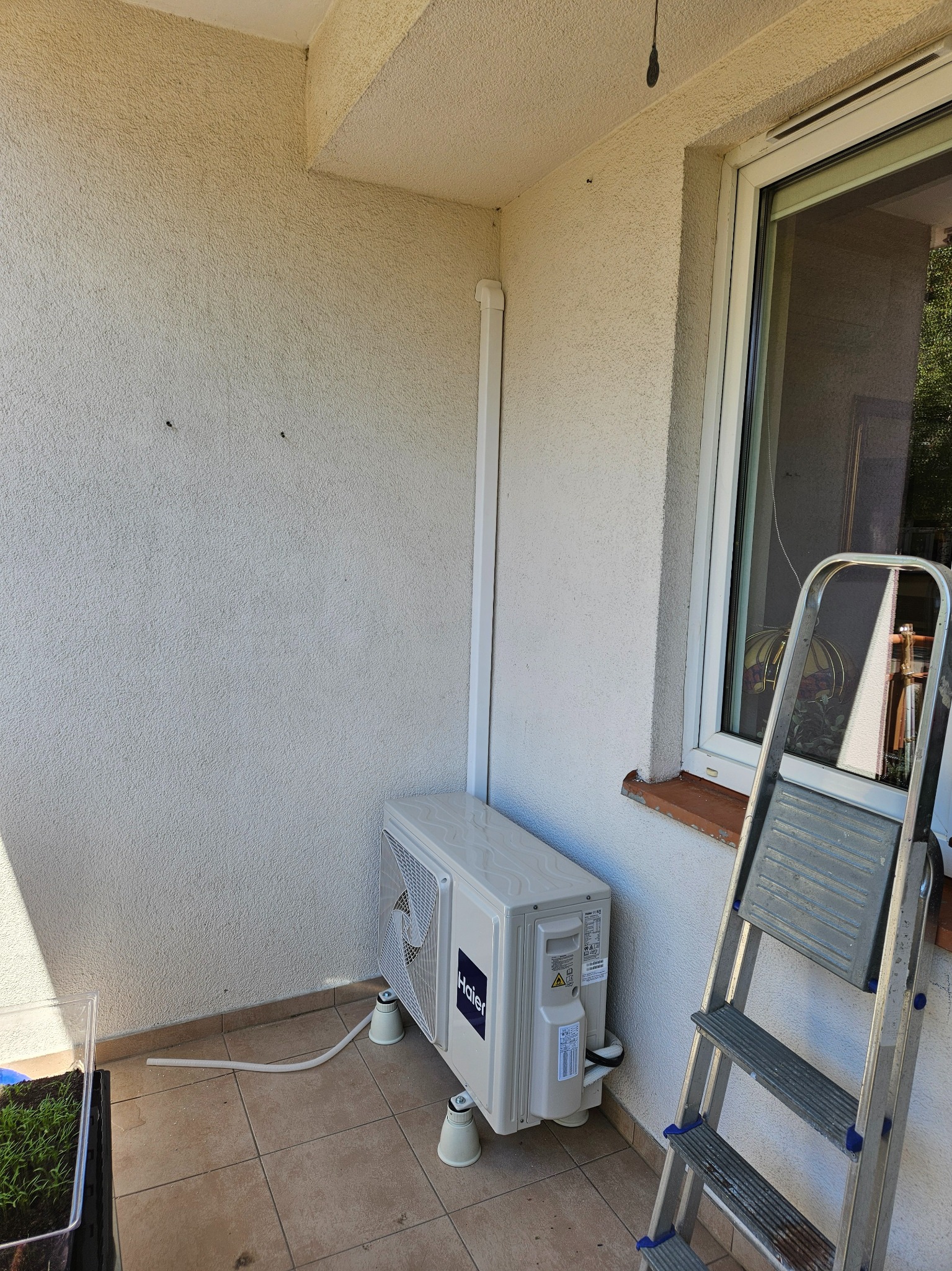 Zewnętrzna jednostka klimatyzatora Haier na balkonie, zamontowana na wspornikach, obok drabina aluminiowa oparta o okno.