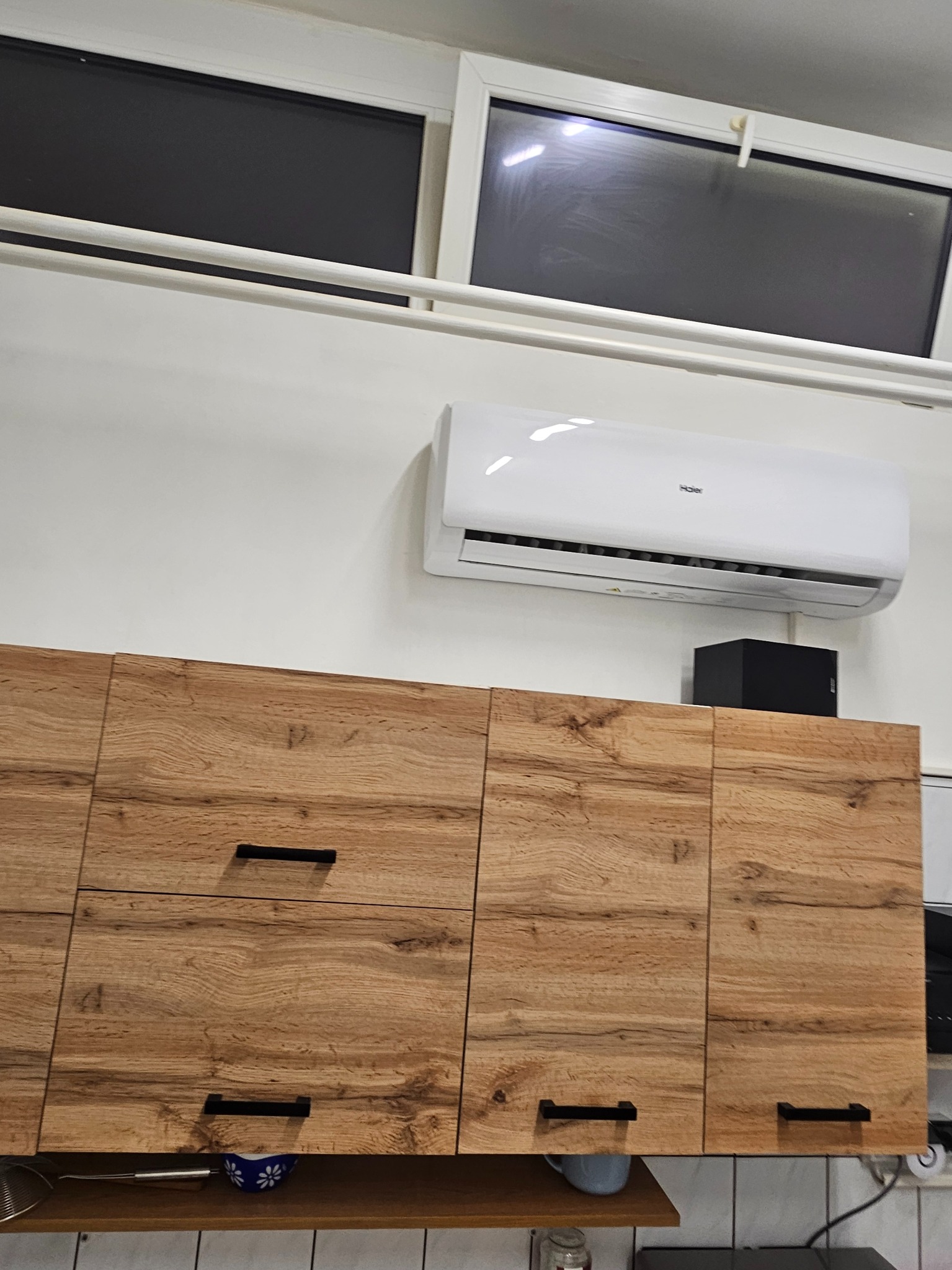 Biały klimatyzator Haier zamontowany nad drewnianymi szafkami kuchennymi z czarnymi uchwytami, widoczne okno z roletami.