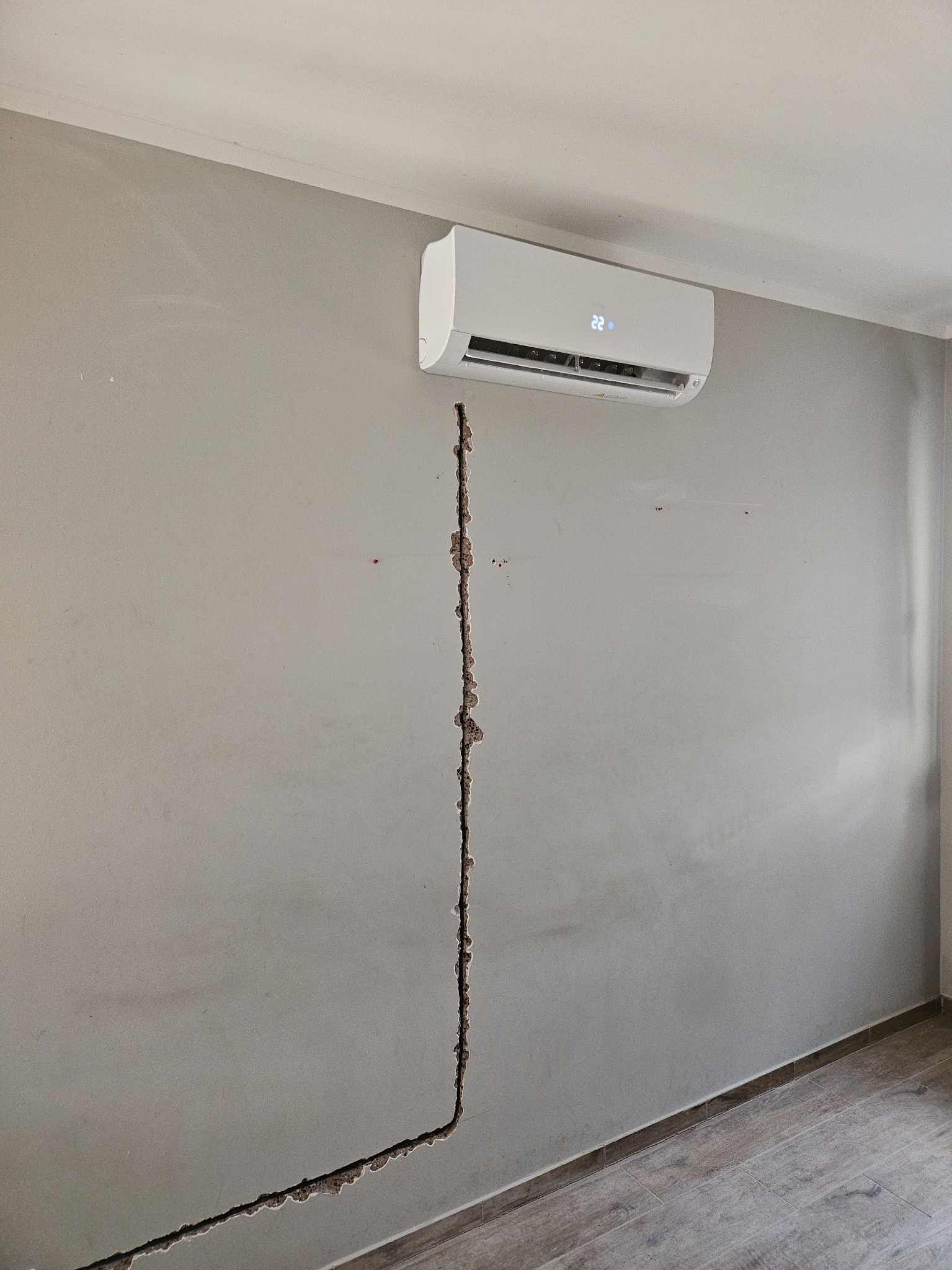 Biały klimatyzator ścienny zamontowany nad wyżłobieniem w szarej ścianie, przez które poprowadzone zostaną instalacje.