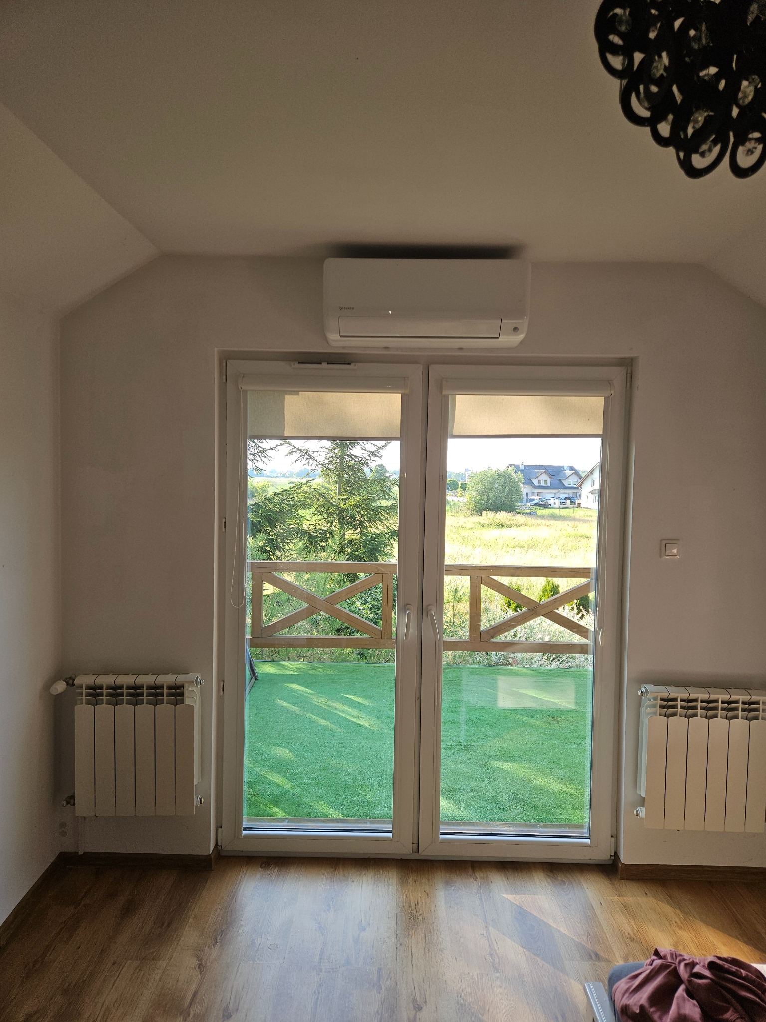 Biały klimatyzator ścienny zamontowany nad dwuskrzydłowymi drzwiami balkonowymi z widokiem na zielony trawnik i domy w oddali, z grzejnikami po bokach i drewnianą podłogą.
