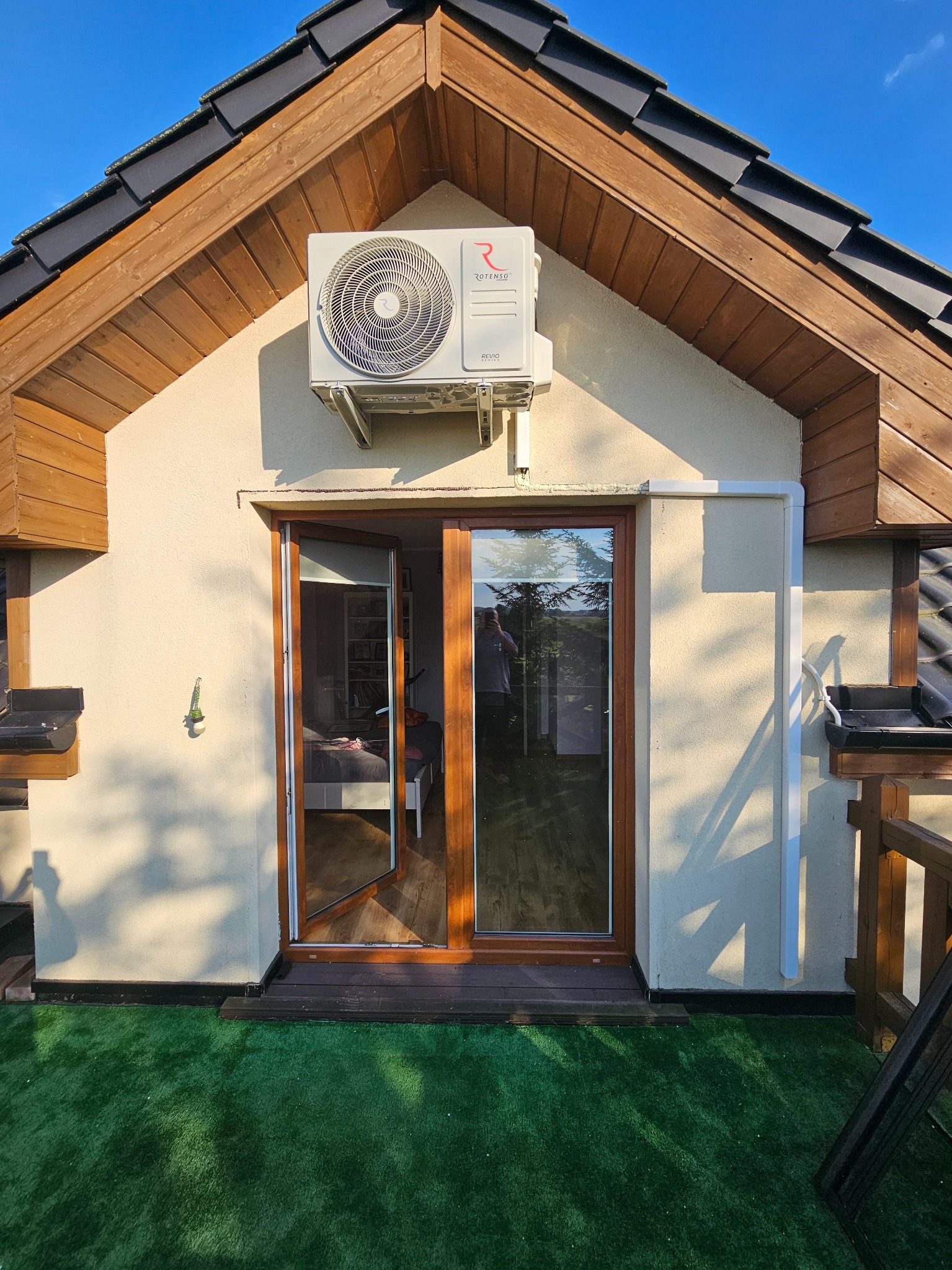 Zewnętrzny agregat klimatyzacji zamontowany nad drzwiami tarasowymi domu z drewnianą podbitką dachową i sztuczną trawą na tarasie.
