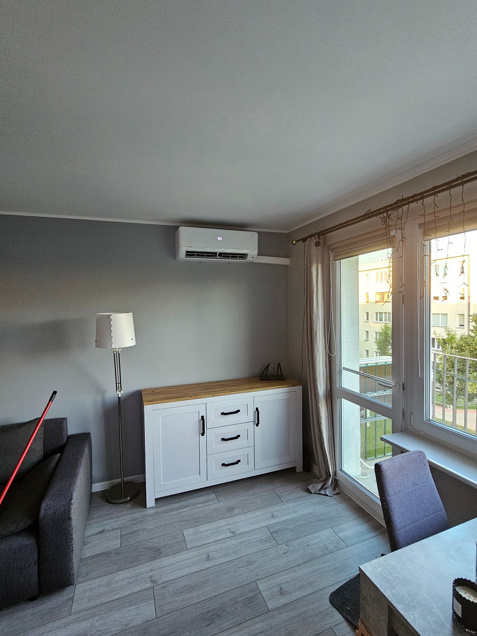 Wnętrze salonu z zamontowanym klimatyzatorem ściennym nad białą komodą z drewnianym blatem, widok na balkon przez okno z beżowymi zasłonami.