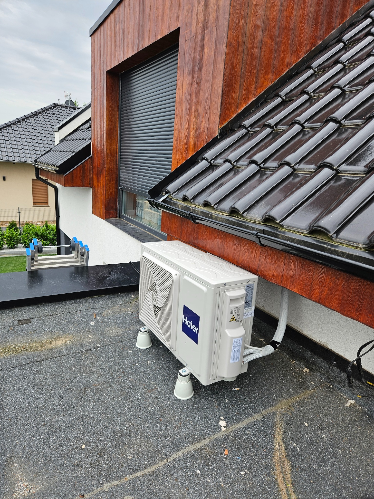 Biała jednostka zewnętrzna klimatyzatora Haier zamontowana na płaskim dachu budynku z drewnianą elewacją i czarną dachówką, widoczne przewody i drabina w tle.