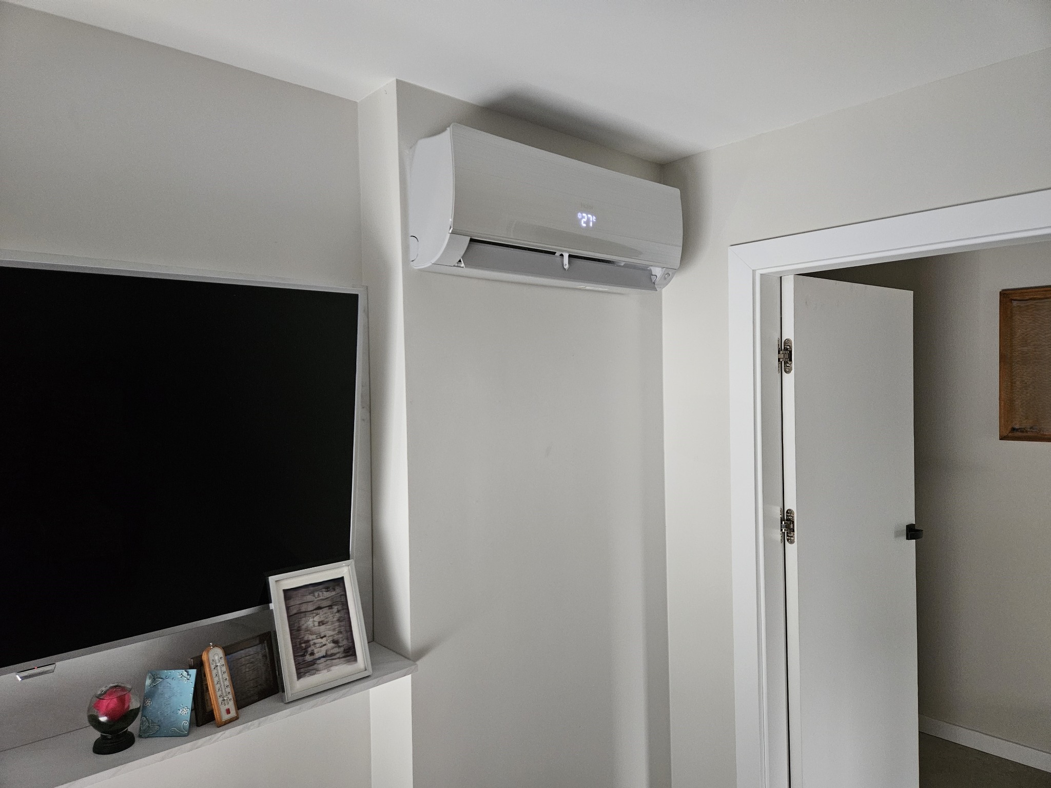 Biały klimatyzator ścienny z wyświetlaną temperaturą 27 stopni Celsjusza, zamontowany w jasnym pokoju obok telewizora i otwartych drzwi.