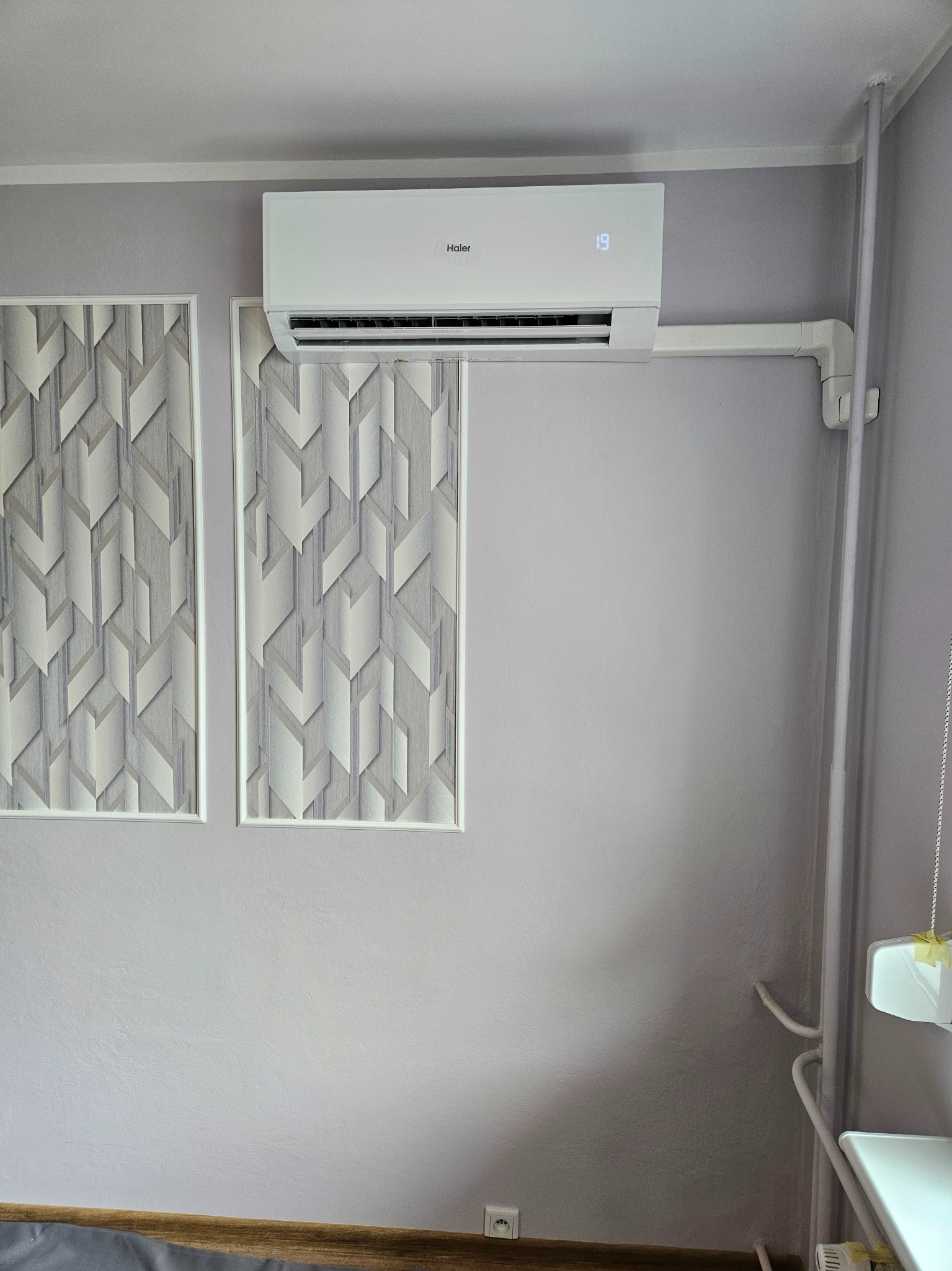 Biały klimatyzator Haier zamontowany na ścianie w pokoju z dekoracyjnymi panelami w geometryczne wzory, widoczne rury instalacji, temperatura ustawiona na 19 stopni.