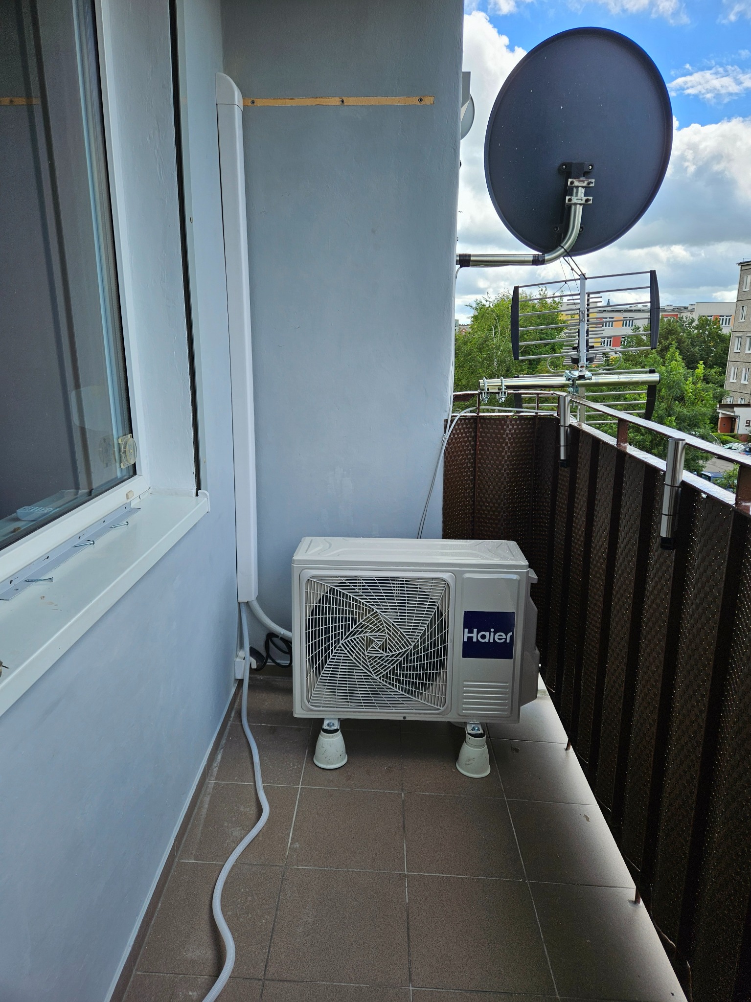 Jednostka zewnętrzna klimatyzatora Haier na balkonie, widoczna antena satelitarna, szare ściany i brązowa balustrada.